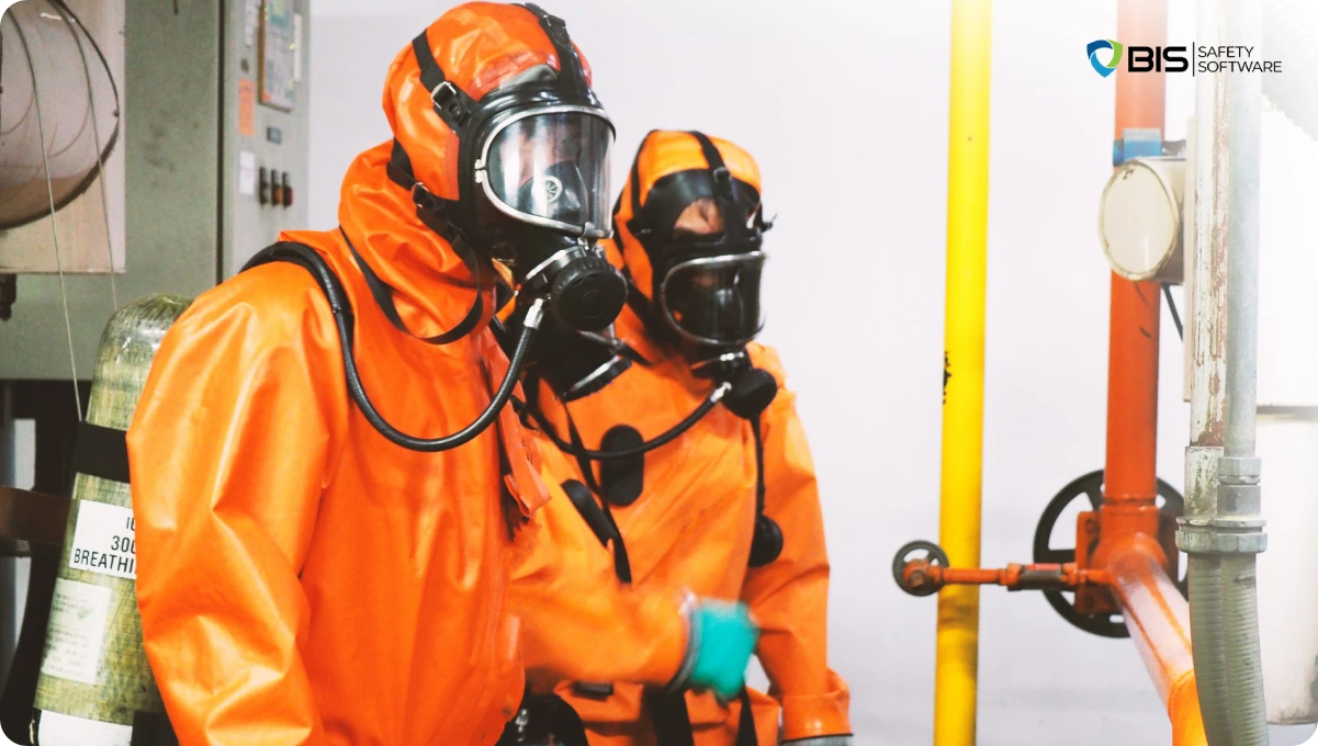 Two workers wearing full-body orange hazmat suits and self-contained breathing apparatus (SCBA) inside an industrial facility, inspecting a pipe system.