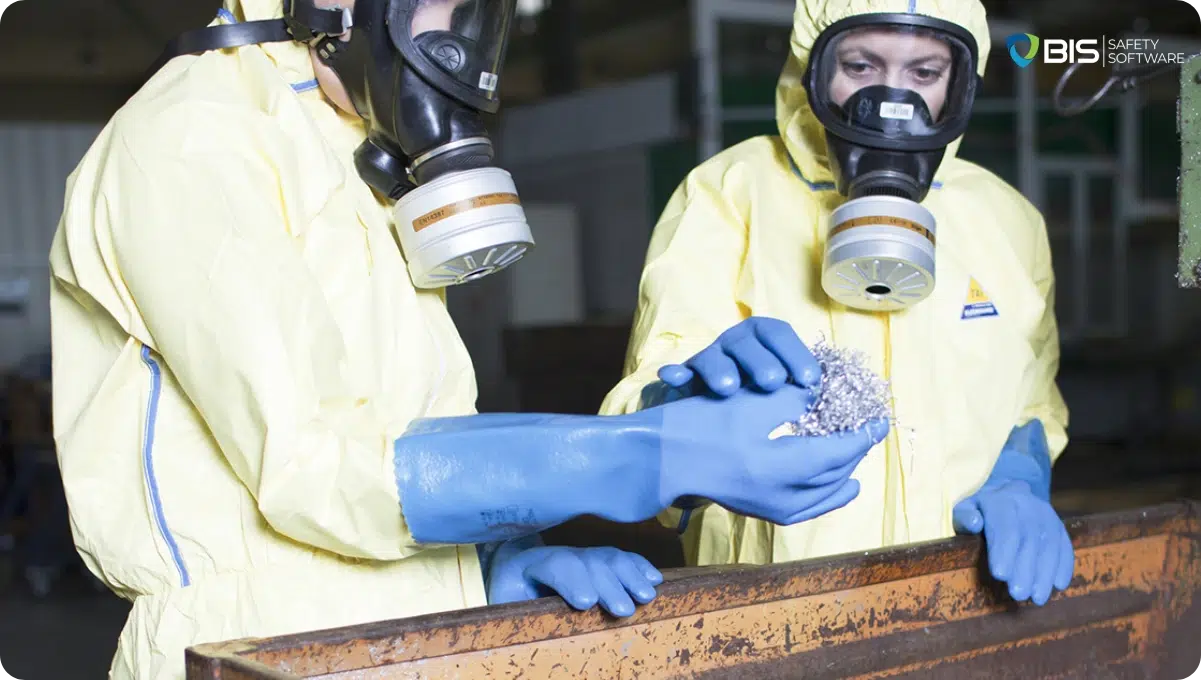 Two workers in yellow hazmat suits, blue chemical-resistant gloves, and full-face respirators examining a handful of metal shavings in an industrial setting.