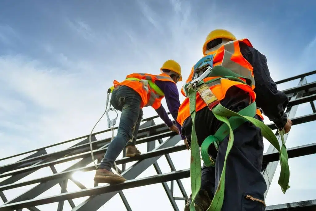 Construction workers in safety gear ascending a steel frame, demonstrating fall protection measures.