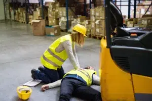 Worker contacting emergency help as another employee administers first aid to an unconscious colleague in a workplace
