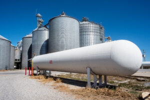 Bulk ammonia storage tank stands at an agricultural facility, with silos and safety bollards in view.