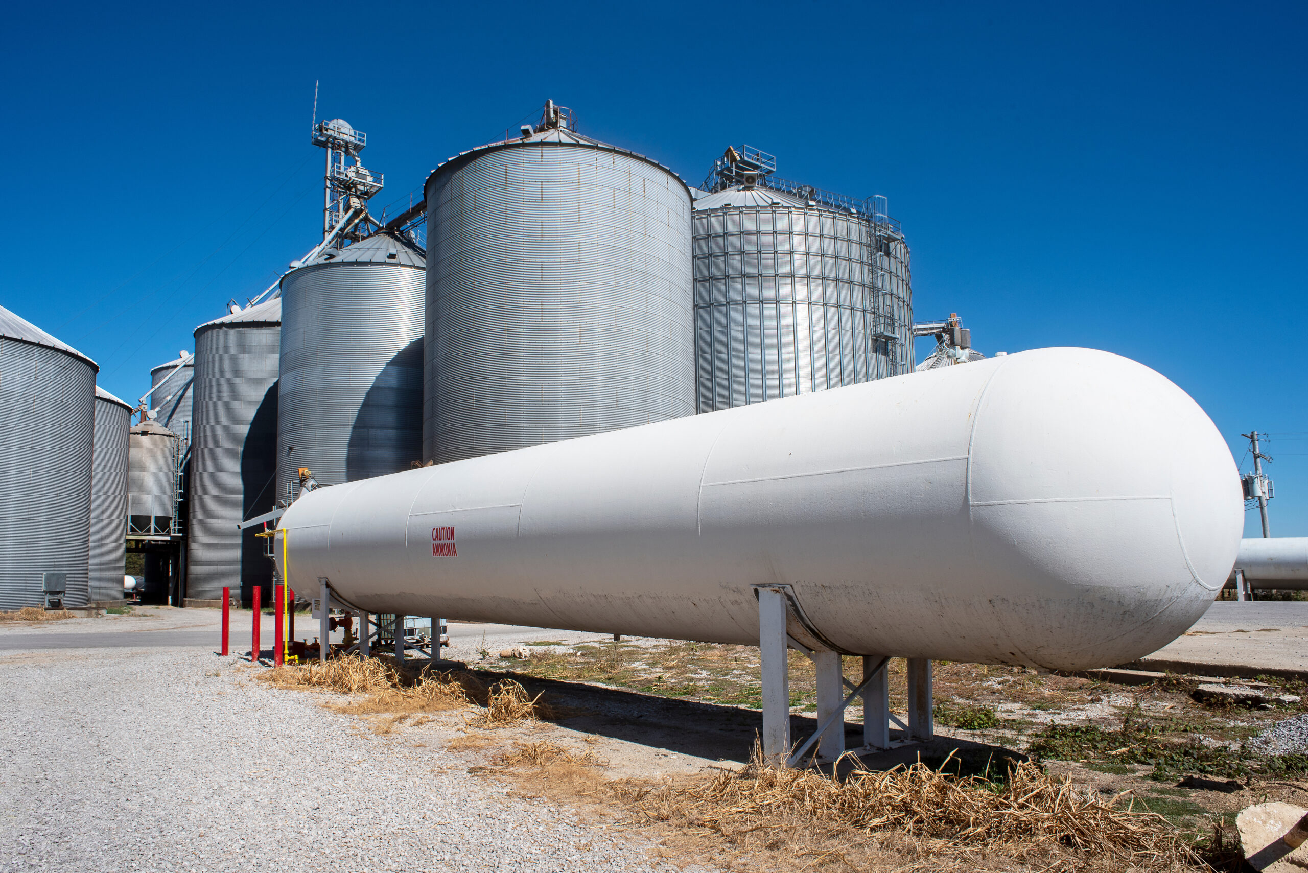 Bulk ammonia storage tank stands at an agricultural facility, with silos and safety bollards in view.