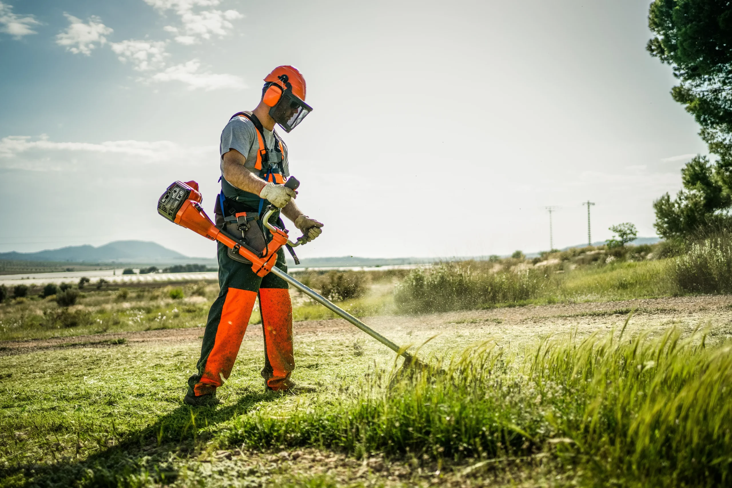 Landscaping worker using a string trimmer while wearing full PPE including visor, gloves, and high-visibility gear.