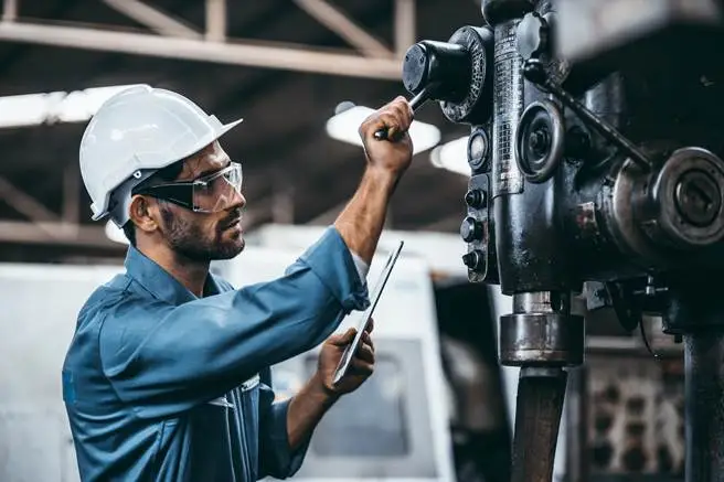 Technician in hard hat inspects industrial machinery while logging notes on a tablet.