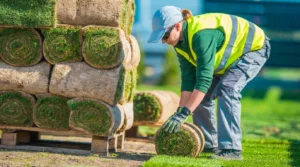 A worker lifting a roll of sod from a pallet while wearing outdoor work clothing.