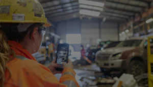 Employee in high-visibility gear capturing a hazard photo on a smartphone in a workshop.